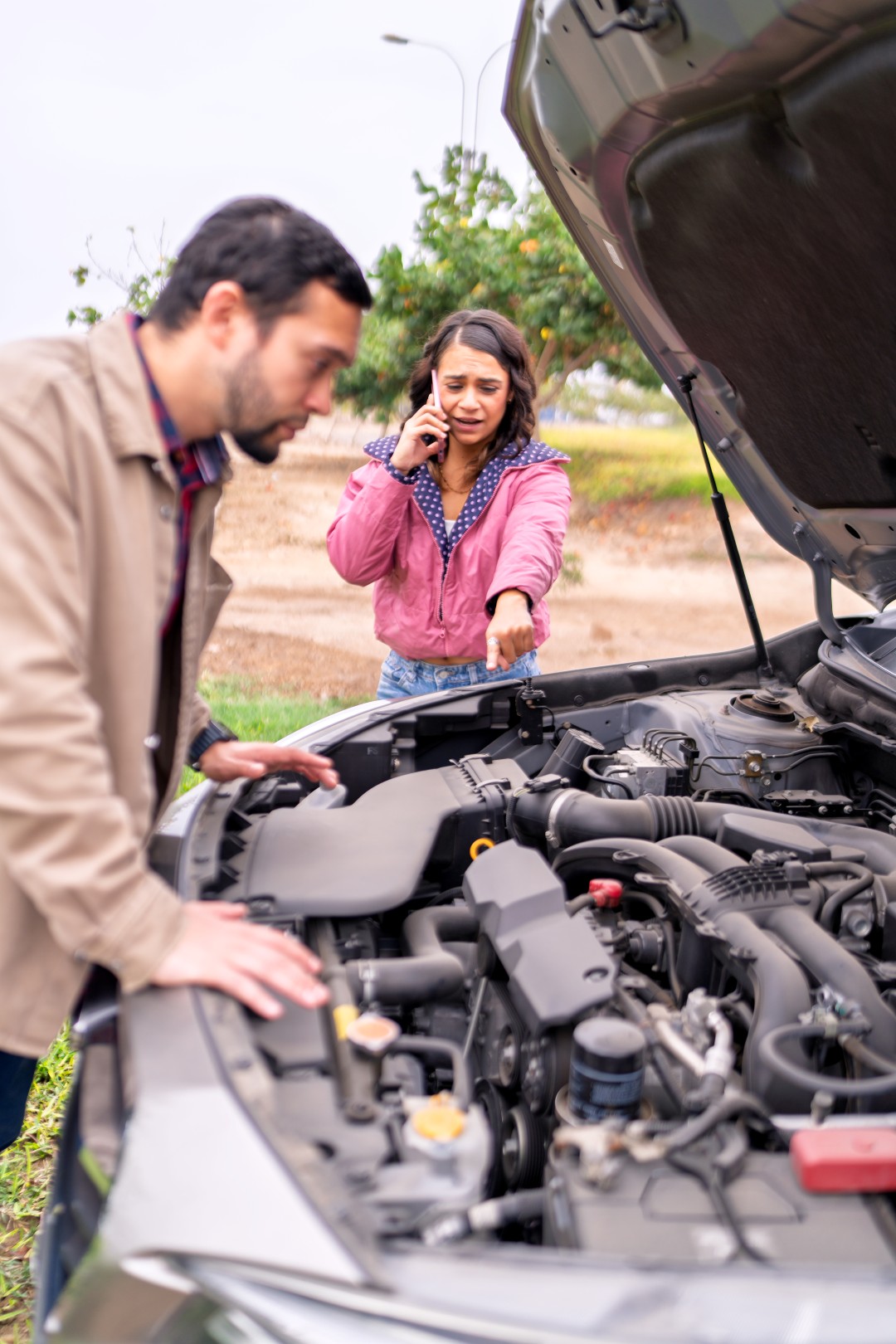 Hispanic couple examining their car's engine after breaking down, woman making phone call for help